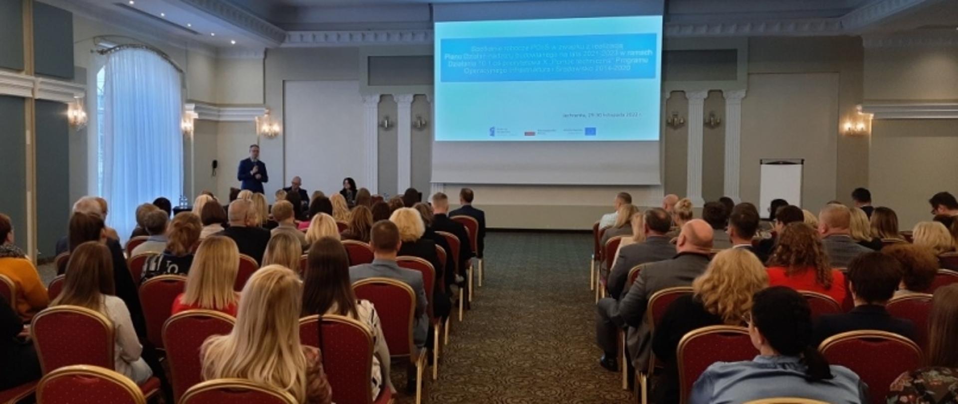 Prelegent podczas spotkania informacyjnego EFS+ w Katowicach; uczestnicy siedzą w sali z czerwonymi krzesłami, ekran z prezentacją w tle.