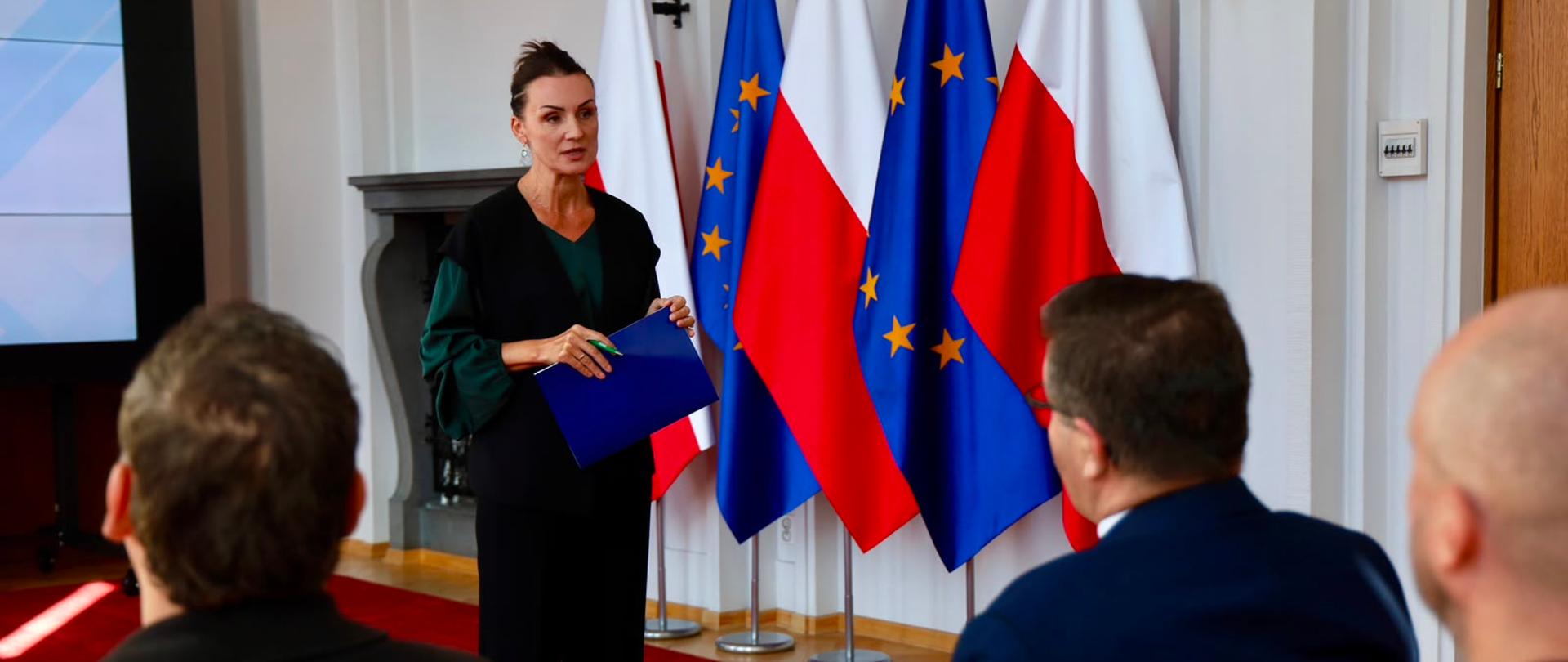 Zdjęcie przedstawia oficjalne spotkanie w pomieszczeniu z czerwonym dywanem na podłodze. Na środku stoi kobieta z ciemnymi włosami spiętymi w kok, ubrana w czarne spodnie, ciemną kamizelkę oraz ciemnozieloną bluzkę z szerokimi rękawami. W rękach trzyma niebieską teczkę i długopis, a jej postawa wskazuje, że przemawia lub prowadzi prezentację. Z prawej strony plecami do kamery siedzi kilku uczestników posiedzenia w eleganckich strojach. W tle, za przemawiającą kobietą, ustawione są na stojakach naprzemiennie flagi Polski (biało-czerwone) oraz Unii Europejskiej (niebieskie z żółtymi gwiazdami). Po lewej stronie widoczny jest fragment dużego ekranu multimedialnego.