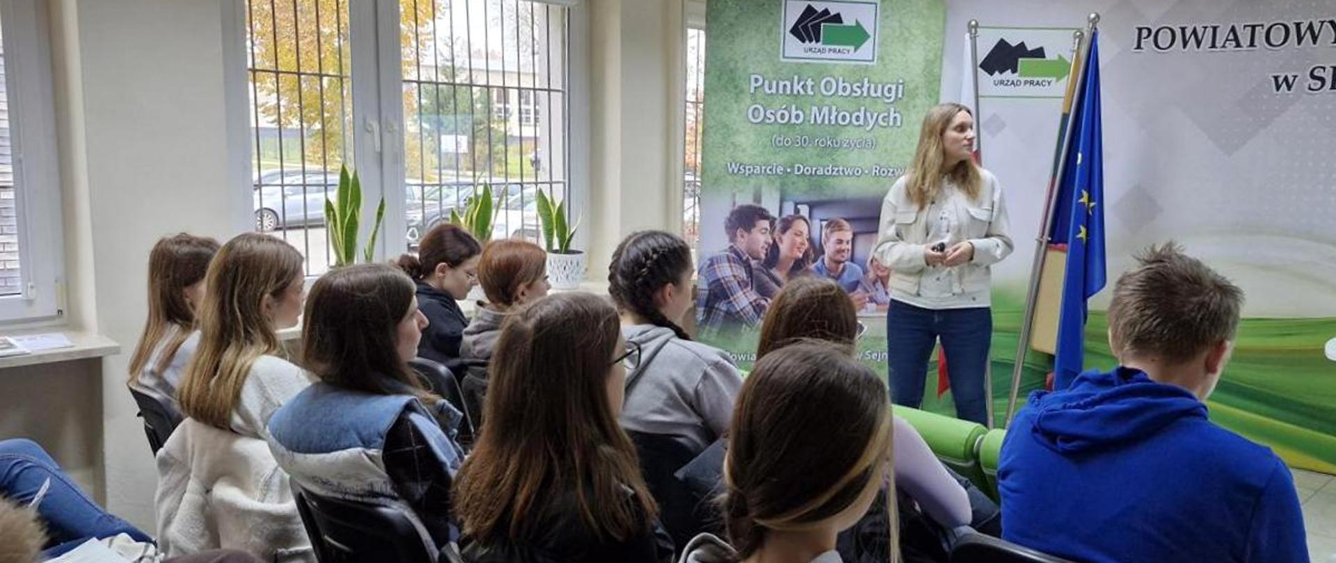 Młodzież słucha prelekcji prowadzonej przez kobietę stojącą na tle banera z logotypem Urzędu Pracy.