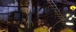 Zdjęcie przedstawia widok w kierunku drewutni i wiaty w których doszło do pożaru. Na trawie i wewnątrz budynków znajdują się spalone elementy wyposażenia oraz drewno. Dodatkowo po prawej stornie na zdjęciu widać przystawioną do dachu drabinę oraz jednego strażaka trzymającego wąż pożarniczy. Strażak ubrany w ubranie specjalne w kolorze ciemnym z elementami odblaskowymi. Na głowie ma żółty hełm. Zdjęcie wykonane w porze nocnej.