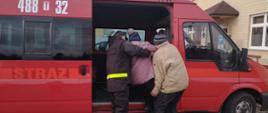 Strażak w czarnym ubraniu koszarowym z żółtymi elementami odblaskowymi wraz z drugim mężczyzną pomagają wsiąść do busa strażackiego kobiecie. 