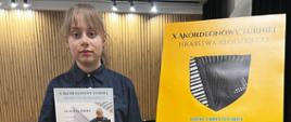 Zdjęcie z konkursu przedstawiające uczennicę trzymającą w jednej ręce dyplom a w drugiej nagrodę w papierowej torbie. Uczennica jest ubrana na czarno i stoi obok sztalugi z plakatem Turnieju Akordeonowego. W tle widać fragment fortepianu i ścianę w lamelach drewnianych. 