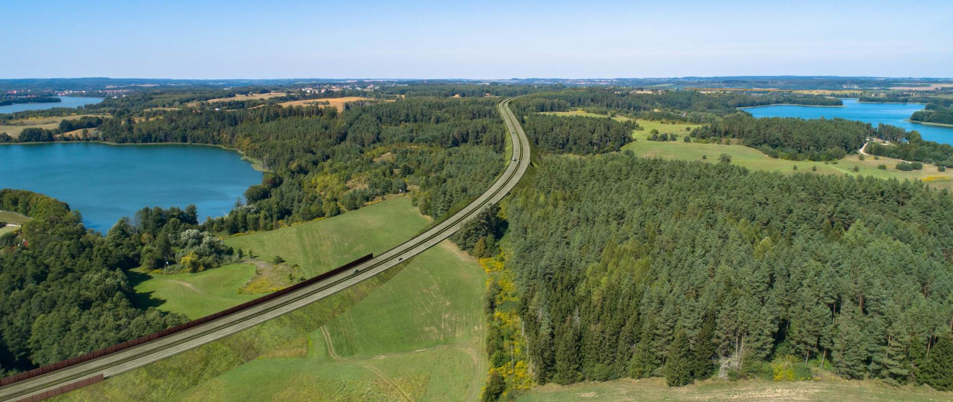 Zdjęcie przedstawia malowniczy krajobraz widziany z lotu ptaka. Przez środek kadru biegnie zakrzywiony tor kolejowy, który wije się przez zielone pola, gęste lasy i w pobliżu kilku jezior. Całość otacza bujna roślinność, a niebo jest bezchmurne i błękitne, co sugeruje słoneczną, pogodną aurę. Kompozycja obrazu ukazuje harmonijne współistnienie infrastruktury drogowej z naturalnym środowiskiem.