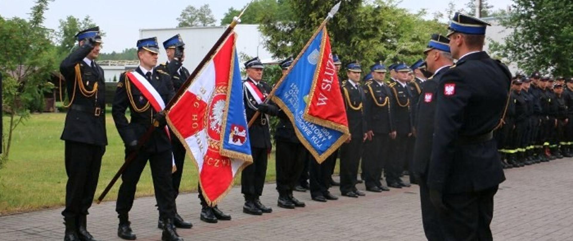 1 Zdjęcie przedstawia moment oddawania honoru poprzez salutowanie zastępcy komendanta wojewódzkiego PSP w Toruniu oraz dowódcę uroczystości. Z lewej strny zdjęcia poczet sztandarowy i pododdział strażaków. W oddali samochody pożarnicze, teren zielony i zabudowania