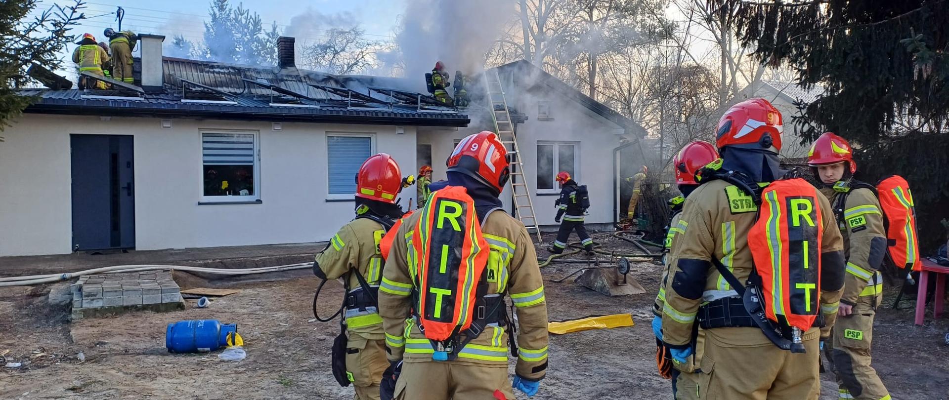 Strażacy w umundurowaniu specjalnym w kolorze żółtym, z aparatami ochrony dróg oddechowych w trakcie działań gaśniczych poddasza budynku rodzinnego.