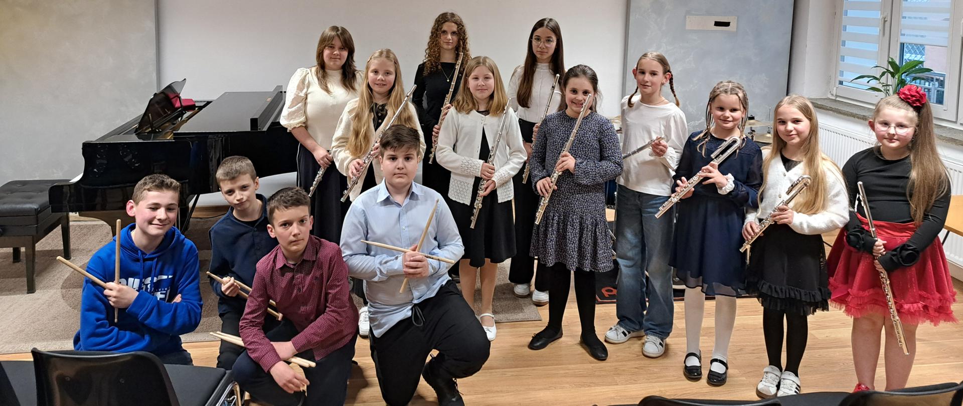 Grupa uczniów szkoły muzycznej stoi w sali kameralnej, pozując do zdjęcia po koncercie. Dzieci trzymają instrumenty – flety poprzeczne oraz pałeczki perkusyjne. Część uczniów stoi w rzędzie, a kilku chłopców kuca z przodu. W tle widoczny jest fortepian oraz jasne ściany z panelami akustycznymi. Uczniowie są elegancko ubrani i uśmiechają się do zdjęcia.