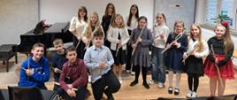Grupa uczniów szkoły muzycznej stoi w sali kameralnej, pozując do zdjęcia po koncercie. Dzieci trzymają instrumenty – flety poprzeczne oraz pałeczki perkusyjne. Część uczniów stoi w rzędzie, a kilku chłopców kuca z przodu. W tle widoczny jest fortepian oraz jasne ściany z panelami akustycznymi. Uczniowie są elegancko ubrani i uśmiechają się do zdjęcia.