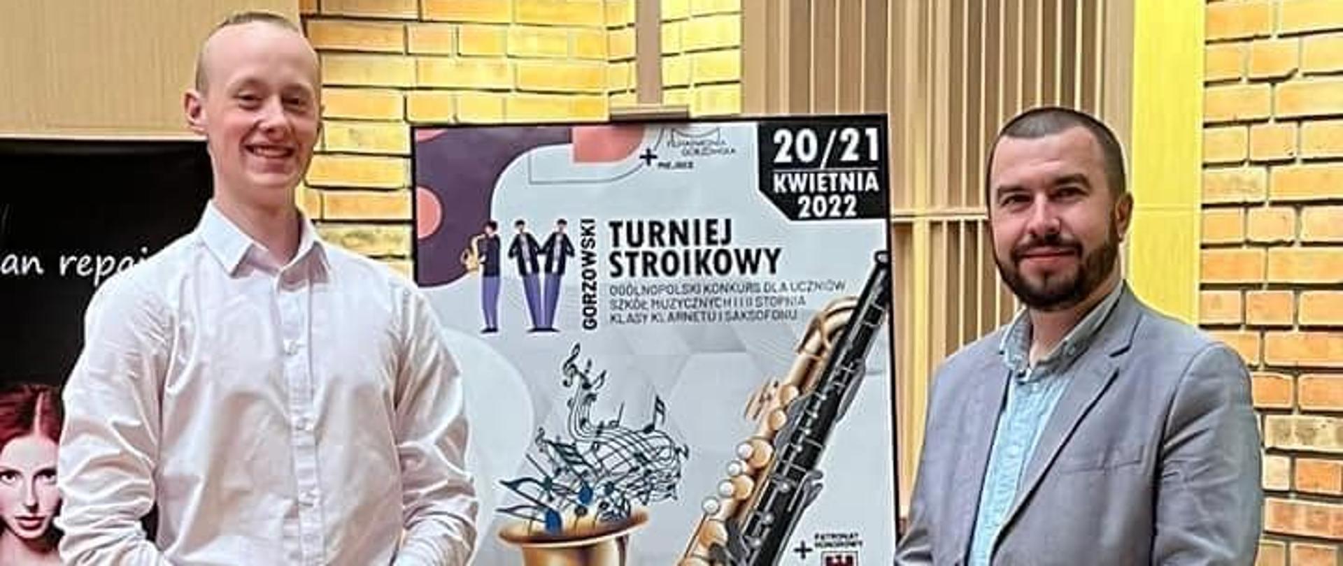 Laureat i Pedagog stoją obok sztalugi z plakatem konkursu, ubrani w stroje oficjalne, w tle mur z cegły.