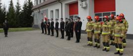 Na zdjęciu widać osiemnastu strażaków, w tym dowódcę uroczystości, poczet sztandarowy oraz pododdział. Dwunastu strażaków ubranych jest w czarne marynarki ze stopniami oraz czapkami z daszkiem a sześciu ubranych jest w żółte stroje i czerwone hełmy. Strażacy stoją w szeregu (oprócz dowódcy uroczystości, który stoi do nich bokiem) na placu wewnętrznym przed budynkiem komendy. Jest widno.