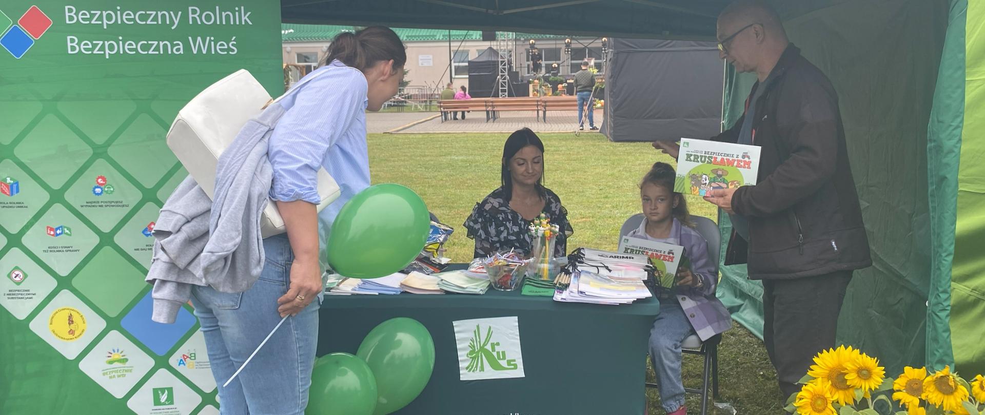 Stoisko KRUS. W centrum zielony namiot opatrzony hasłem: „Pracuj Bezpiecznie”. Po lewej baner z logiem Kasy, obok kobieta sięgająca po broszury prewencyjne. Pod namiotem stolik z broszurami oraz materiałami prewencyjnymi. Za stolikiem siedząca pracownica KRUS, a przy niej siedzące dziecko, które trzyma w ręku grę prewencyjną. Po prawej stronie stoi mężczyzna także trzymający w rękach grę prewencyjną.