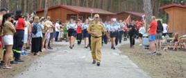 Teren ośrodka wypoczynkowego. Betonową drogą pomiędzy drewnianymi budynkami strażak w mundurze koloru piaskowego maszeruje strażak, za nim maszeruje orkiestra dęta i obozowicze. Po obu stronach drogi znajduje sie liczne grono gości, którzy przyglądają się maszerującym. 