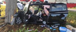 Zdjęcie przedstawia wypadek w miejscowości Redlino w powiecie białogardzkim. Zdjęcie przedstawia czarny pojazd marki Ford B, który przodem od strony kierowcy uderzył w drzewo. Za wrakiem pojazdu samochody strażackie