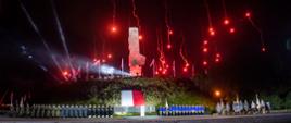 Nad pomnikiem spadają czerwone race pod pomnikiem stoją żołnierze harcerze oraz umieszczona jest flaga Polski.