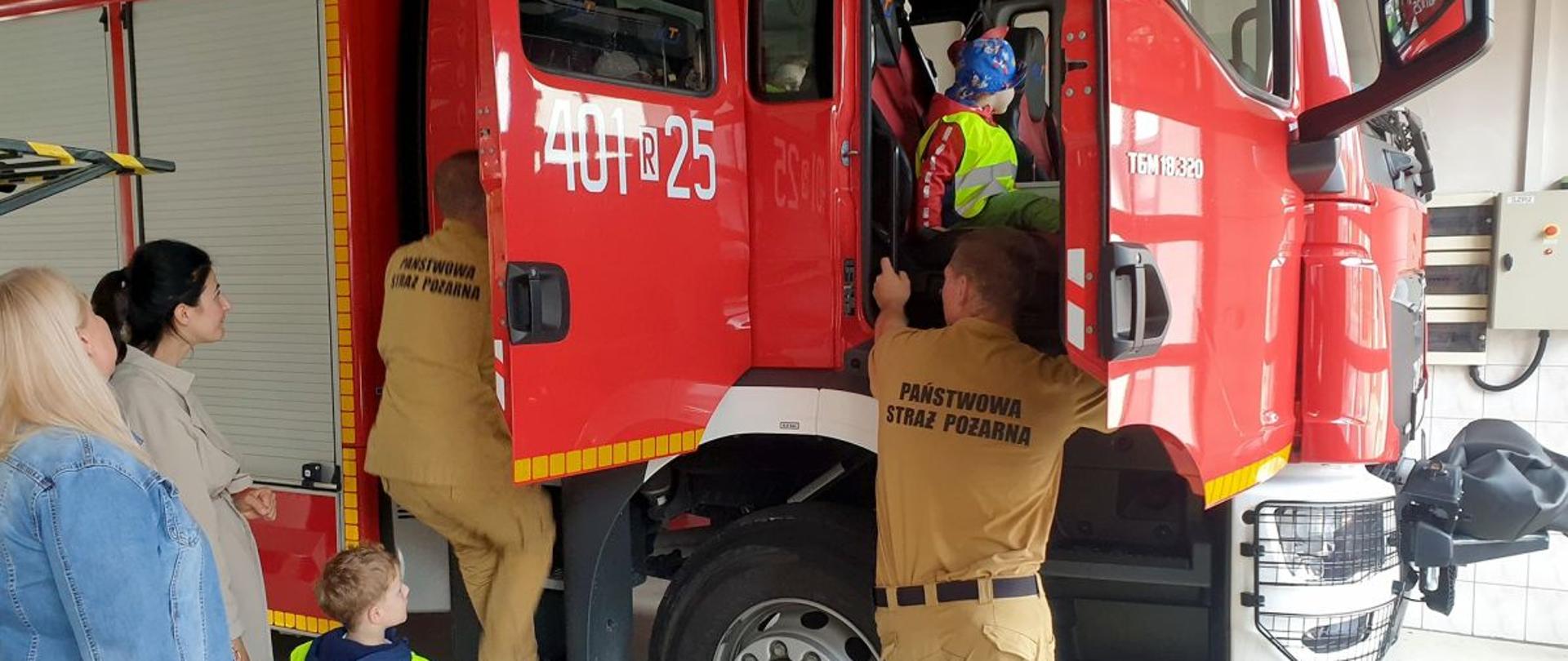 Zdjęcie przedstawia garaż jednostki ratowniczo-gaśniczej w której przy samochodzie gaśniczym stoi dwóch strażaków, w aucie siedzi jedno dziecko a przy samochodzie stoi drugie a obok niego dwie Panie nauczycielki. 