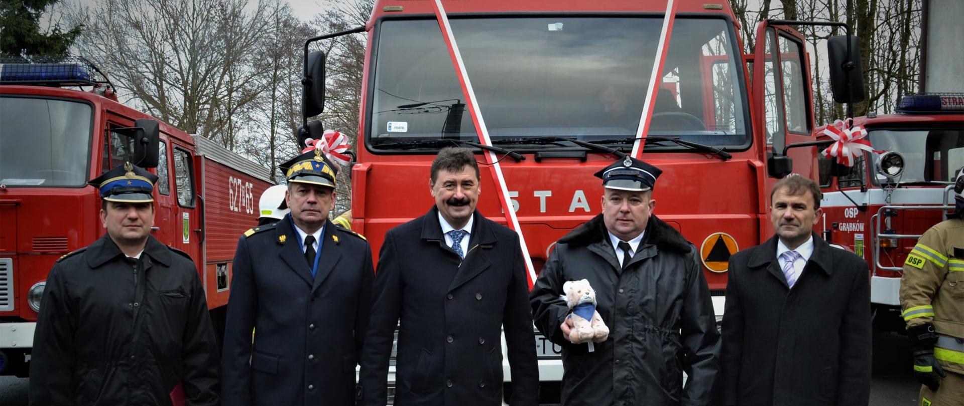 Zaproszeni na uroczystość goście , wśród nich zastępca komendanta głównego PSP oraz druh OSP pozują do zdjęcia przed przekazywanym dla OSP samochodem, po prawo i lewo ustawione dwa inne samochody gaśnicze