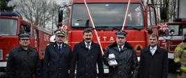 Zaproszeni na uroczystość goście , wśród nich zastępca komendanta głównego PSP oraz druh OSP pozują do zdjęcia przed przekazywanym dla OSP samochodem, po prawo i lewo ustawione dwa inne samochody gaśnicze