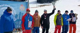 Siedmiu mężczyzn ubranych w zimowe ubrania stoi za podium, sześciu ma zawieszone medale na szyi, jeden trzyma w górze puchar 