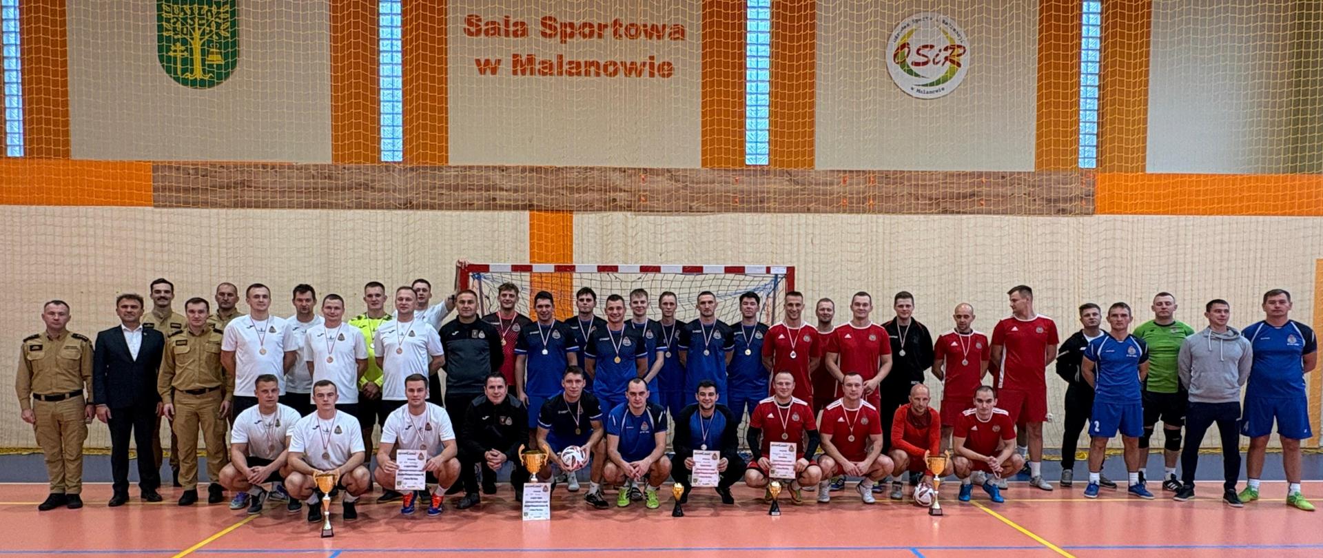 Pamiątkowe, zbiorowe zdjęcie grupowe wszystkich uczestników turnieju wykonane w hali sportowej. Na pomarańczowym parkiecie stoi oraz klęczy w kilku rzędach kilkudziesięciu mężczyzn – w tym drużyny w białych, granatowych, czerwonych i niebieskich strojach sportowych, w większości z medalami na szyjach. Przed klęczącymi zawodnikami ustawione są puchary i dyplomy.
Na skraju po lewej stronie stoją oficjele: dwaj strażacy w piaskowych mundurach oraz mężczyzna w garniturze. Na dużej, beżowo-pomarańczowej ścianie w tle widnieje wyraźny napis „Sala Sportowa w Malanowie”, obok którego umieszczono herb gminy oraz logo ośrodka sportu.