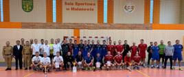 Pamiątkowe, zbiorowe zdjęcie grupowe wszystkich uczestników turnieju wykonane w hali sportowej. Na pomarańczowym parkiecie stoi oraz klęczy w kilku rzędach kilkudziesięciu mężczyzn – w tym drużyny w białych, granatowych, czerwonych i niebieskich strojach sportowych, w większości z medalami na szyjach. Przed klęczącymi zawodnikami ustawione są puchary i dyplomy.
Na skraju po lewej stronie stoją oficjele: dwaj strażacy w piaskowych mundurach oraz mężczyzna w garniturze. Na dużej, beżowo-pomarańczowej ścianie w tle widnieje wyraźny napis „Sala Sportowa w Malanowie”, obok którego umieszczono herb gminy oraz logo ośrodka sportu.