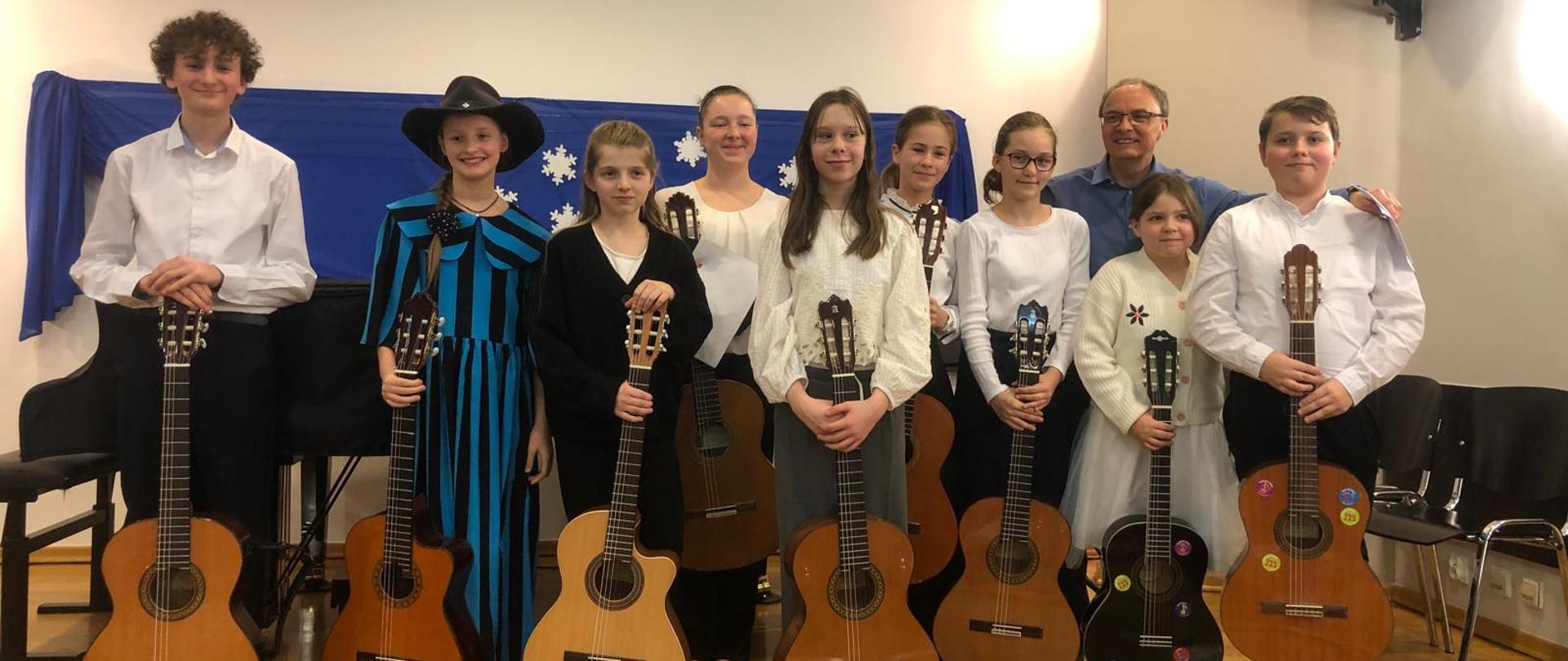 Zdjęcie zostało wykonane podczas koncertu uczniów klasy gitary. Na zdjęciu grupa odświętnie ubranych uczniów. Wszyscy z nich trzymają gitarę. Razem z uczniami na zdjęciu mężczyzna z niebieskiej koszuli i okularach. W tle za nimi czarny fortepian, a na ścianie niebieski materiał i białymi śnieżynkami.
