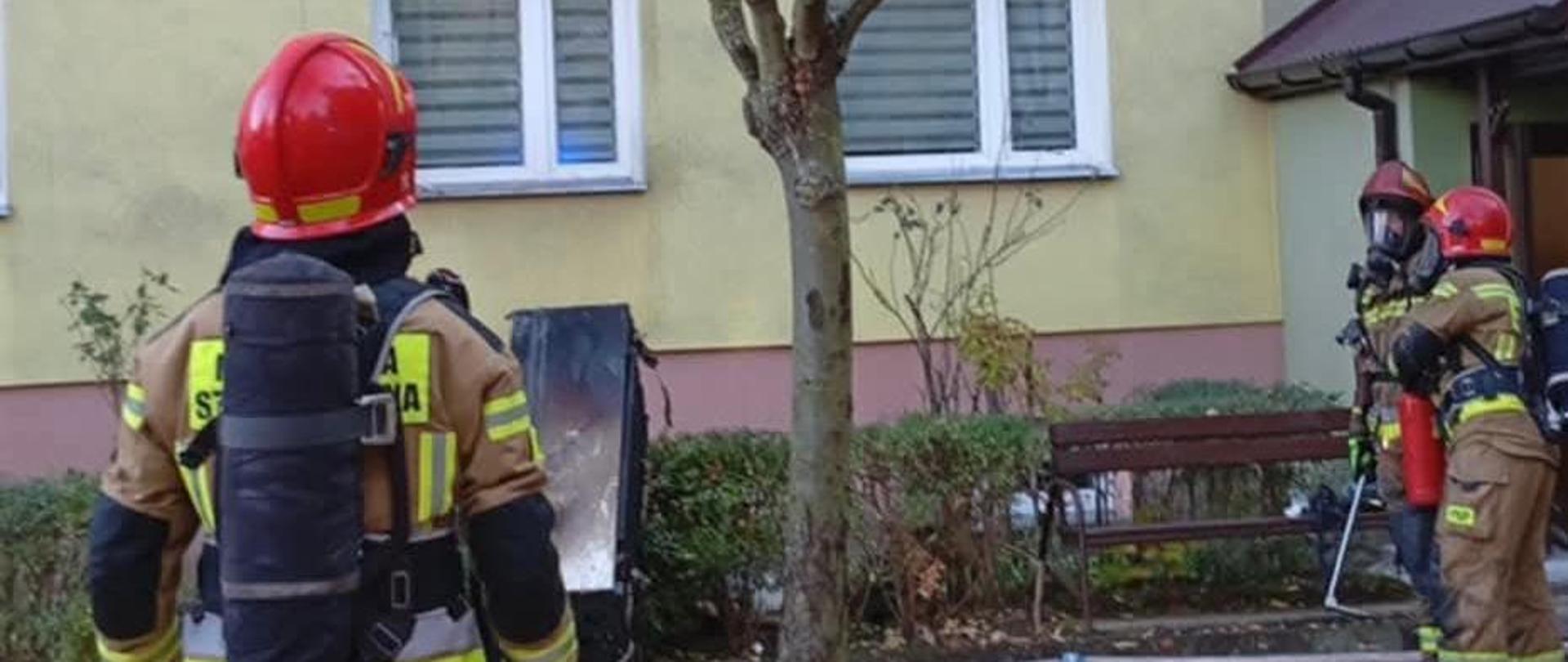 Na zdjęciu widoczni strażacy wyposażeniu w aparaty ochrony układu oddechowego na tle widoczny budynek mieszkalny w którym doszło do pożaru mieszkania.