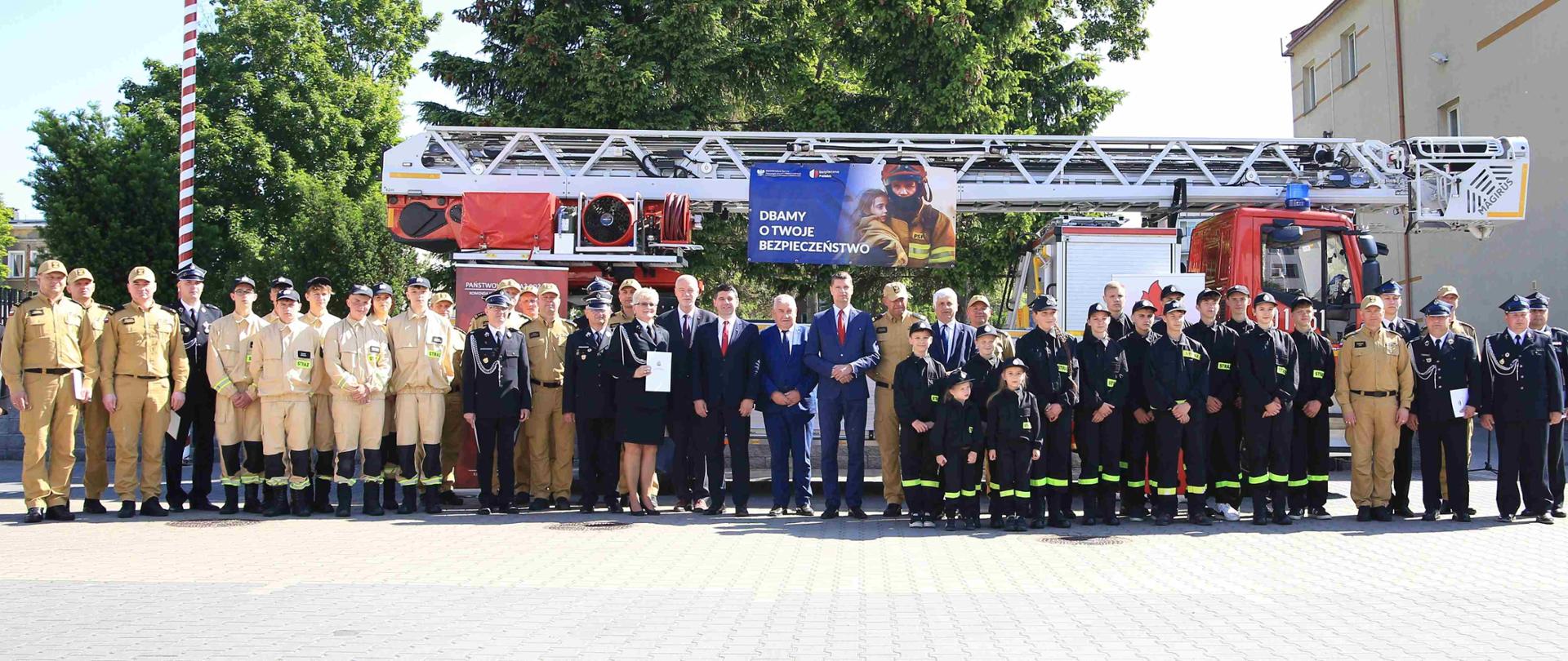 Na tle strażackiej drabiny w dwóch rzędach stoi kilkadzieisąt osób większość to strażacy w piaskowych i granatowych mundurach. 