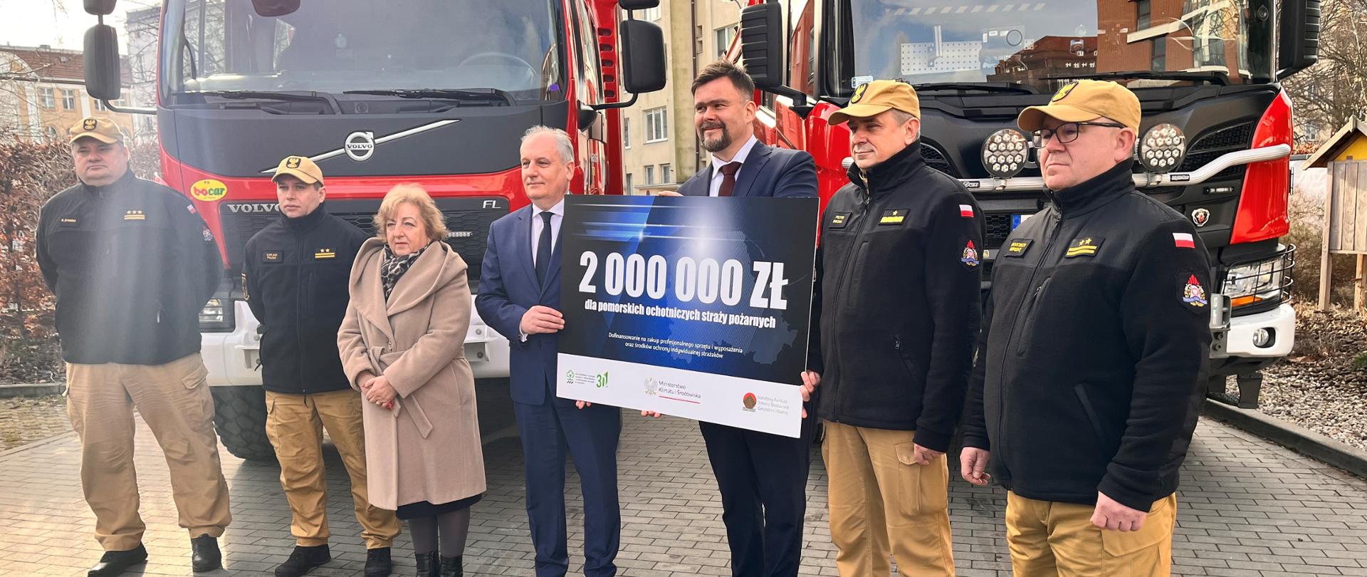 Strażacy oraz osoby cywilne mężczyźni oraz jedna kobieta stoją przed samochodami gaśniczymi dwóch mężczyzn trzyma tablicę z liczbą dwa miliony złotych. 