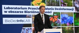 Na zdjęciu widać mężczyznę który wypowiada się przy mikrofonie. W tle na ekranie widać prezentację z napisem "Laboratorium przeciwdziałania informacji w obszarze bioróżnorodności. BioDezInfoLab".