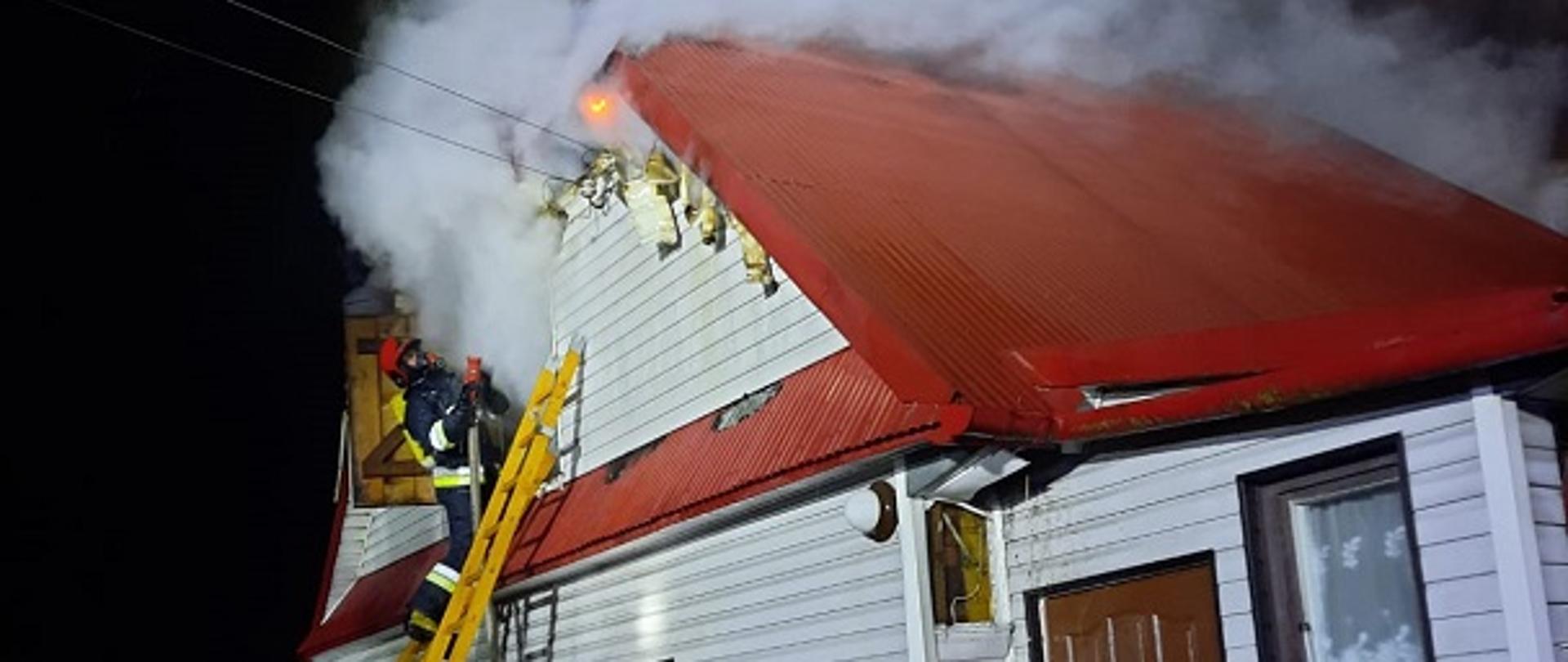 Zdjęcie obrazuje drewniany budynek parterowy. Z poddasza budynku wydobywa się duża ilość dymu. Na drabinie opartej o budynek znajduje się strażak ubrany w aparat oddechowy, który gasi palące się poddasze przez otwór rewizyjny. Na dole drabina jest asekurowana przez dwóch strażaków.