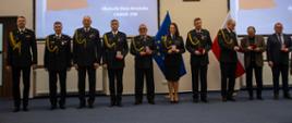 Grupowe zdjęcie wyróżnionych – osoby odznaczone medalami stoją w szeregu na tle slajdu z nazwą wydarzenia.