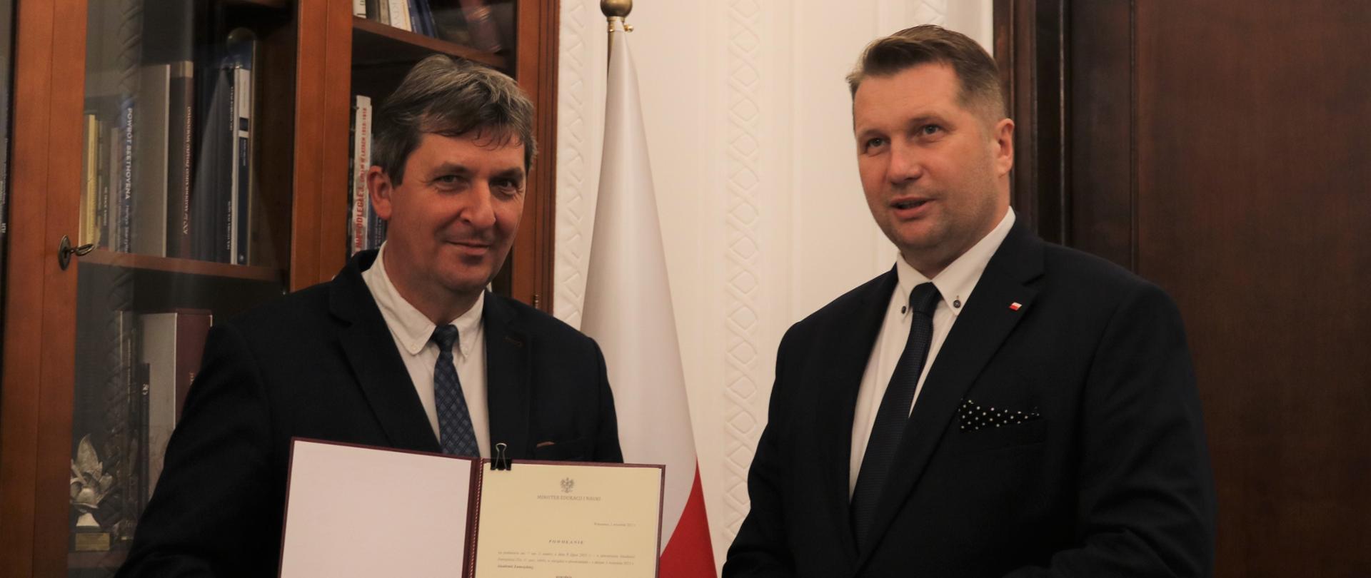 Doktor habilitowany Paweł Skrzydlewski trzyma w ręku teczkę z nominacją na stanowisko rektora Akademii Zamojskiej. Jest ubrany w garnitur, białą koszulę i krawat. Obok stoi minister Przemysław Czarnek ubrany w garnitur, białą koszulę i krawat. W butonierce ma czarną poszetkę w białe groszki. Za Skrzydlewskim gablota z książkami i biało-czerwona flaga.