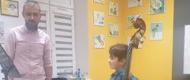 Na zdjęciu znajdują się pedagog prowadzący warsztaty - Wojciech Front oraz uczeń z kontrabasem w sali rytmicznej PSM w kolorach szaro, żółty, biały, na ścianach wiszą kolorowe obrazki muzyczne
