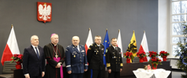 Przedstawiciele służb mundurowych podległych Ministerstwu Spraw Wewnętrznych i Administracji biskup oraz wojewoda pomorski stoją obok siebie za nimi stoją flagi Polski Unii Europejskiej województwa pomorskiego niedaleko stoi stolik z białym obrusem świecą oraz kiatem gwiazdą betlejemską. 