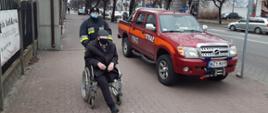 Strażak prowadzi po chodniku wózek inwalidzki, na którym siedzi starszy mężczyzna ubrany na czarno. Obok stoi samochód strażacki.