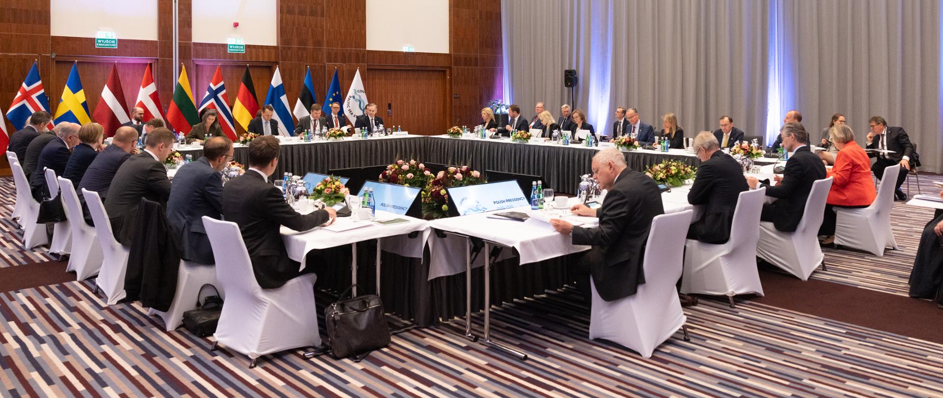 A group of people is sitting at tables arranged in a square. They have white chairs. The tables are covered with white tablecloths. There are flags in the background. 