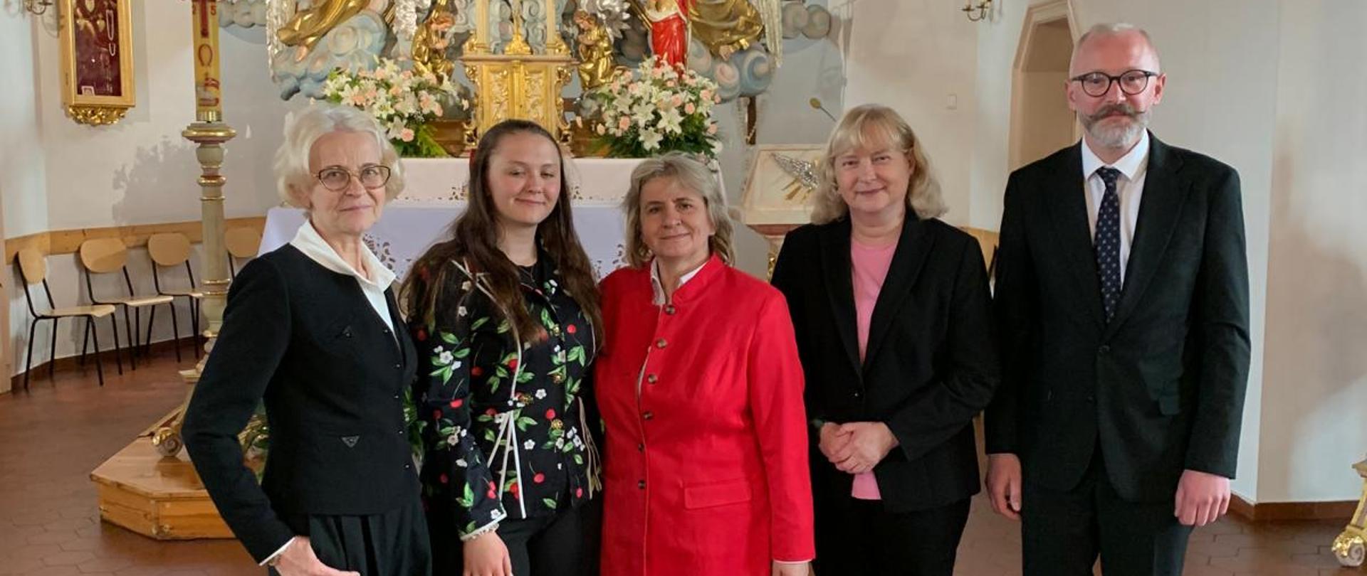 Kolorowe zdjęcie w kościele w Raszczycach w jednym rzędzie stoi 5 osób: dyplomantka Anna i jej nauczyciele . Wszyscy ubrani w galowe stroje z uśmiechami na twarzach. 