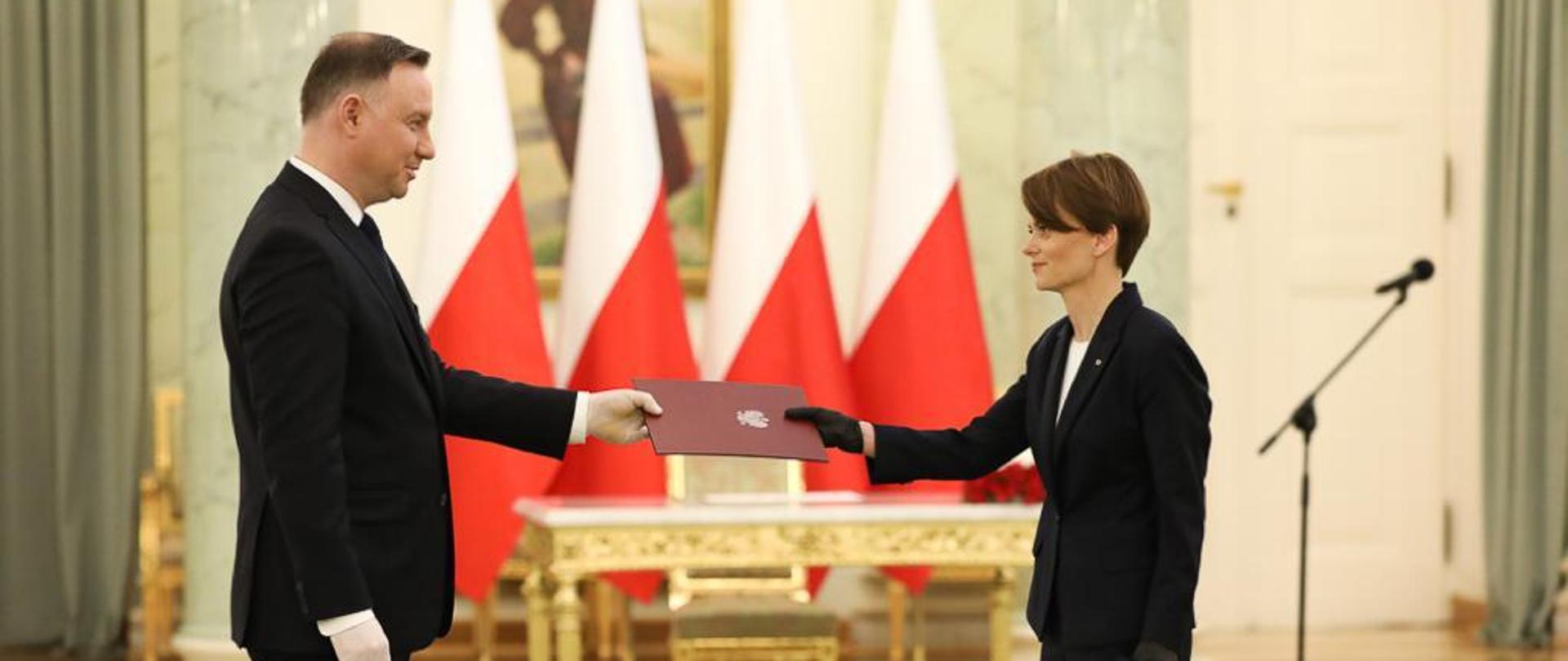 Prezydent Andrzej Duda wręcza minister Jadwidze Emilewicz teczkę. W tle elegancki stół, krzesło oraz polskie flagi.