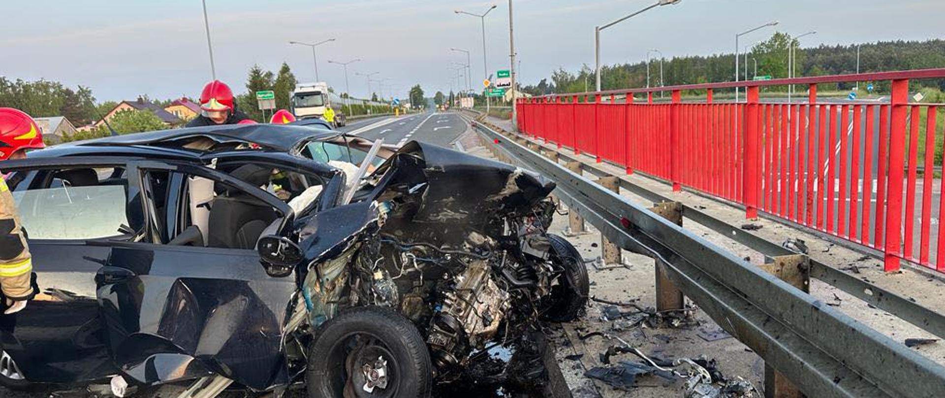 Na pierwszym planie roztrzaskany Fiat i strażacy pracujący przy aucie.