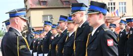 Na pierwszym planie oficer w ciemnym mundurze galowym z naszywką „Państwowa Straż Pożarna” na ramieniu podaje rękę strażakowi stojącemu w szeregu. Szereg składa się z funkcjonariuszy i funkcjonariuszek w galowych mundurach z czapkami rogatywkami, którzy trzymają w dłoniach białe dokumenty. W tle widoczne są fragmenty zabytkowych kamienic oraz flagi na budynku Urzędu Miasta.
