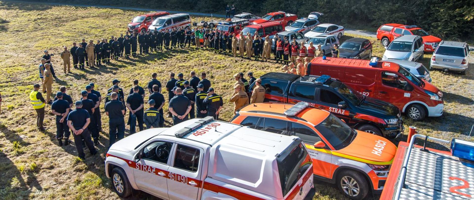 Grupowe zdjęcie podczas przywitania uczestników ćwiczeń i obserwatorów w Jarnołtówku. Za osobami znajdują się samochody straży pożarnej z Polski i Czech.