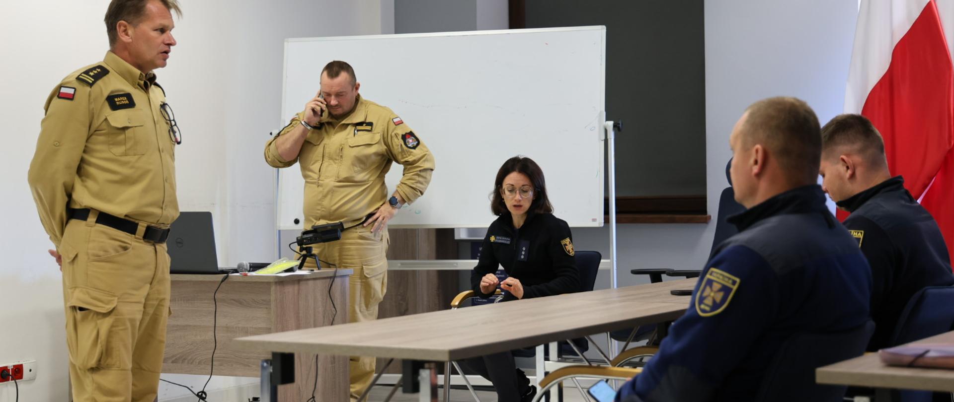 Dwóch strażaków w mundurach służbowych polskich, w pozycji stojącej, zaś obok widok na siedzących trzech strażaków ukraińskich. Trwa rozmowa miedzy nimi. Jeden z polskich strażaków rozmawia przez telefon. W tle wyposażenie sali w postaci tablicy, ławek wykładowych, wyposażenia multimedialnego.