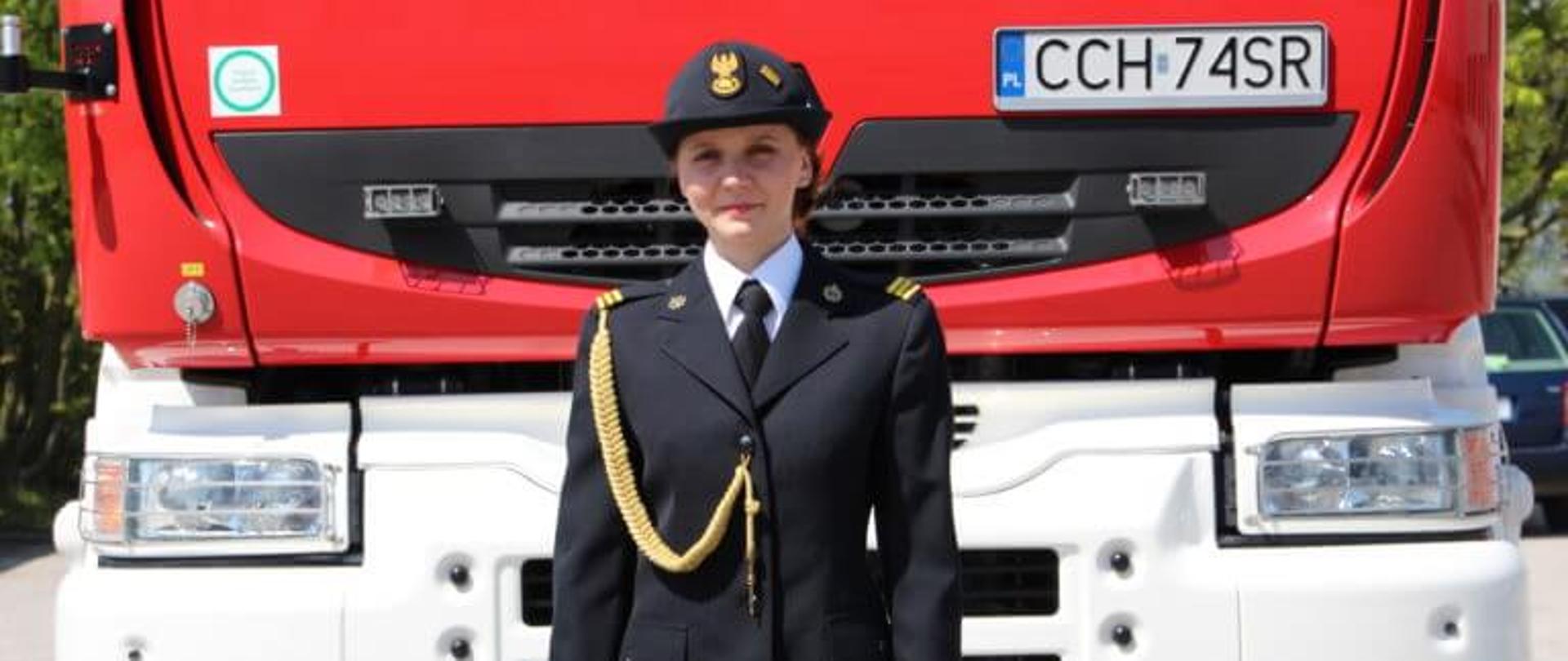 Na zdjęciu stoi strażaczka pozująca do zdjęcia przed wozem bojowym