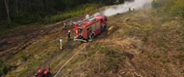Widok z drona na pojazd gaśniczy. Widok na tył auta z niższej wysokości.W tle unosie się dym. Przy pojeździe strażacy.
