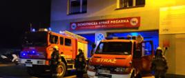 Strażacy w mundurach bojowych stoją przy samochodach pożarniczych wystawionych przed bramami garażowymi. Nad autami na ścianie napis Ochotnicza Straż Pożarna Żory - Rój. Sceneria nocna. 