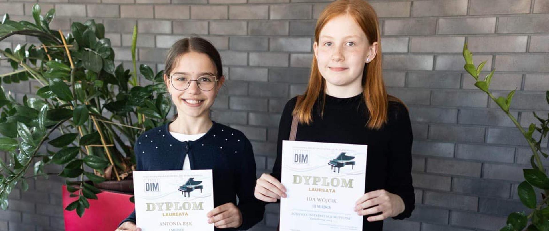 Dwie uśmiechnięte uczennice trzymające dyplomy laureatów na tle szarej, ceglanej ściany. Z tyłu widać kwiaty w czerwonych doniczkach.