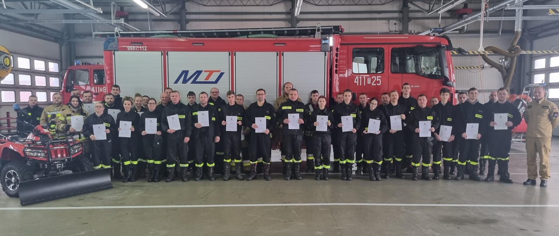 Zdjęcie przedstawia strażaków w umundurowaniu koszarowym na garażu komendy powiatowej PSP w Jędrzejowie. Strażacy trzymają w rękach zaświadczenia. Zdjęcie zrobione na tle wozu strażackiego.