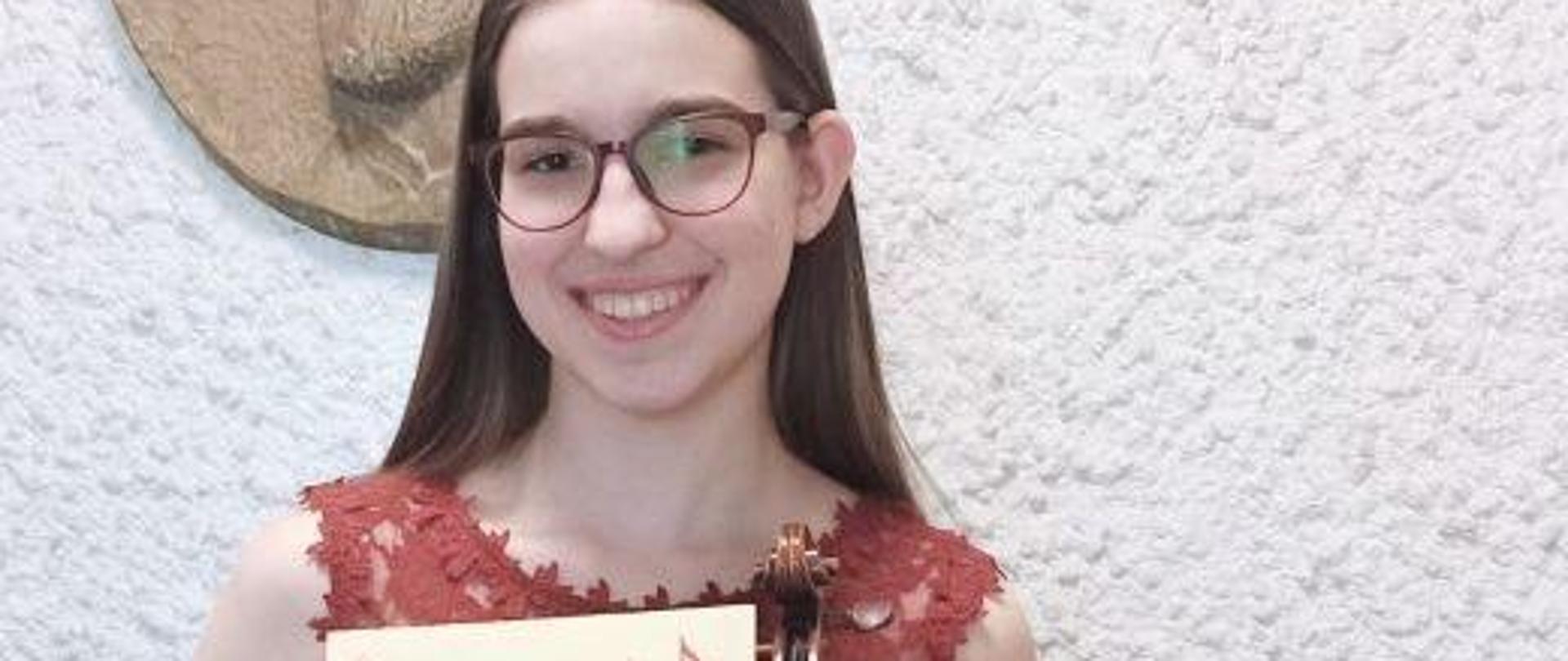 Zdjęcie kolorowe. Dziewczyna ze skrzypcami w koncertowej sukni koloru ceglastego trzyma w rękach dyplom. W tle wizerunek Mieczysława Karłowicza w mosiądzu.