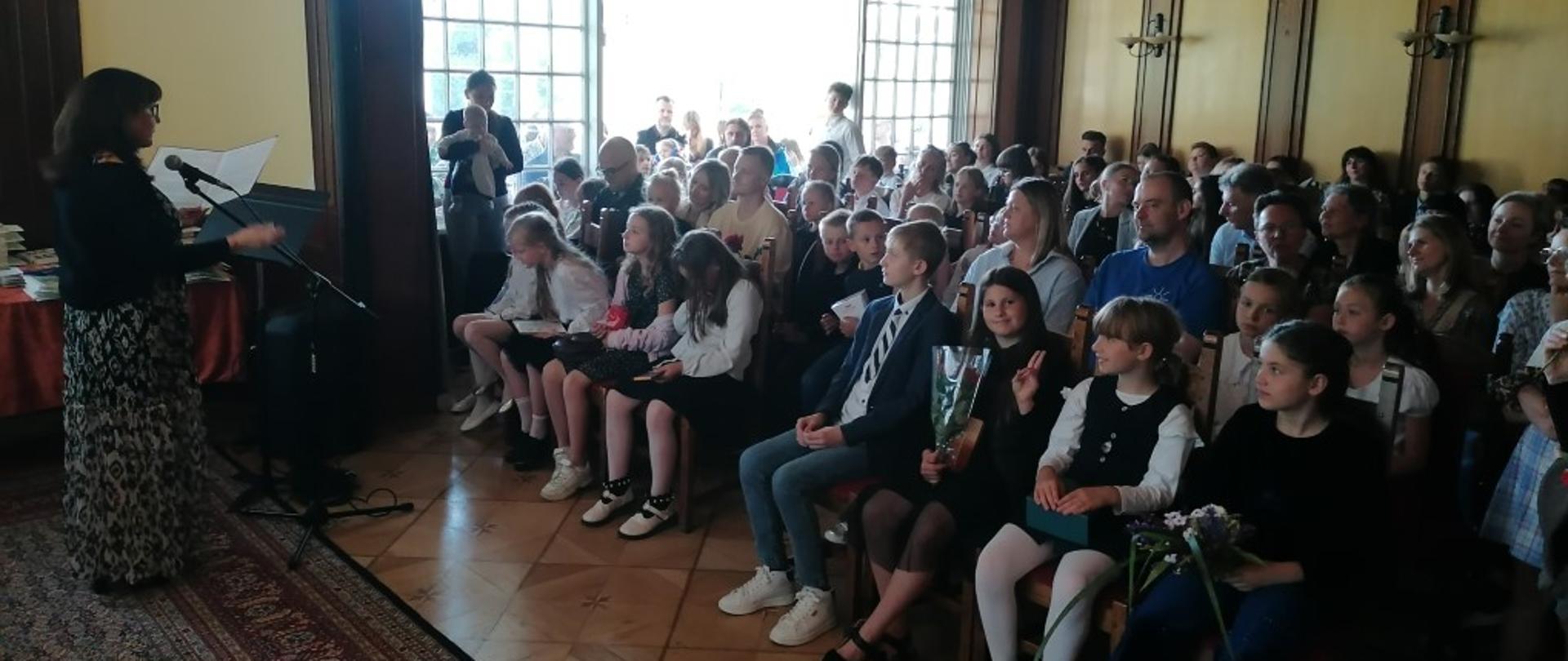 Zdjęcie kolorowe. Kobieta ubrana w czarną bluzkę i kwiecistą spódnicę stoi po lewej stronie, mówiąc do mikrofonu i gestykulując. Przed nią stoi pulpit z nutami lub książką. Po prawej stronie siedzi i stoi duża grupa ludzi, w tym dzieci i dorośli, zwróconych w stronę kobiety. W tle widoczne są duże okna, przez które wpada światło. Pomieszczenie ma drewniane panele na ścianach i ozdobny żyrandol.