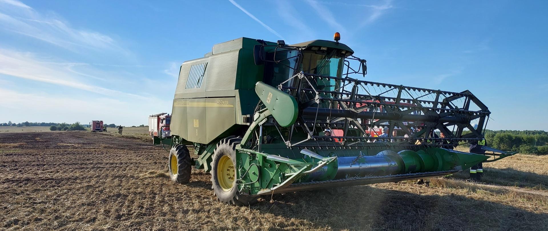 Zdjęcie przedstawia kombajn do prac rolniczych, w którym doszło do zwarcia instalacji elektrycznej. W dalszej perspektywie widać dwa wozy pożarnicze użyte podczas gaszenia ścierniska.