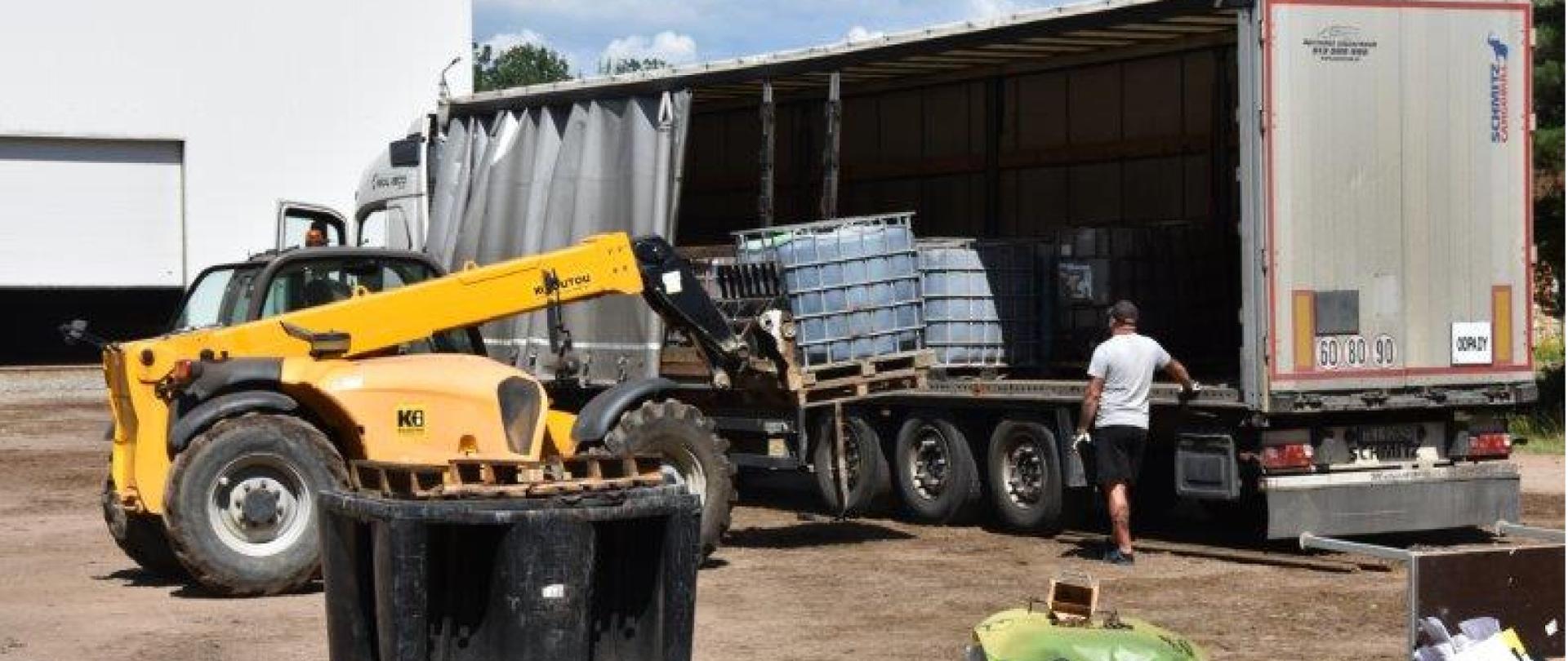 Wózek widłowy ładuje pojemnik typu mauzer na naczepę samochodu ciężarowego. W jej wnętrzu widać załadowane uprzednio inne pojemniki typu mauzer.